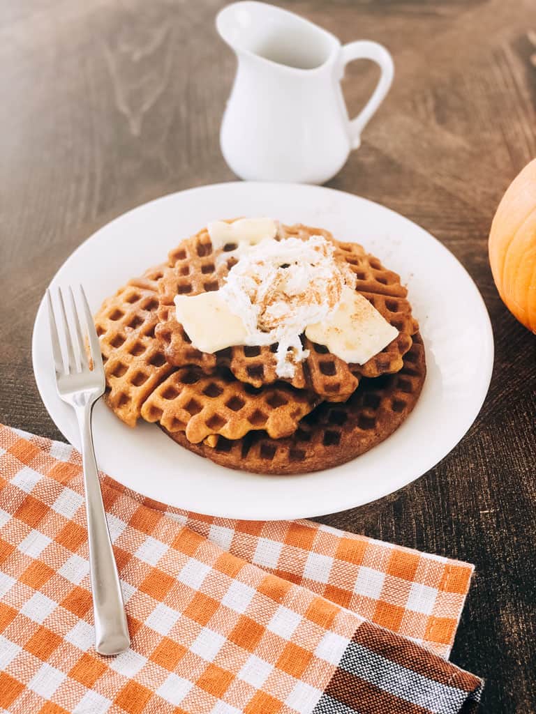 3 pumpkin sourdough waffles stacked on plate and topped with butter, syrup, and whipped cream