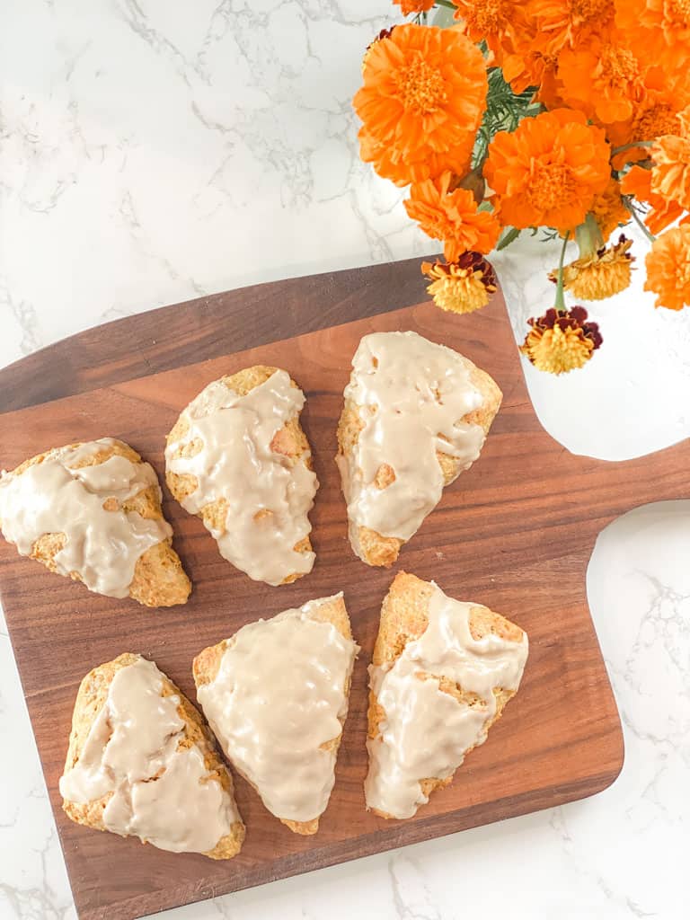 six pumpkin sourdough scones drizzled with maple glaze on a bread board near a vase of orange marigolds