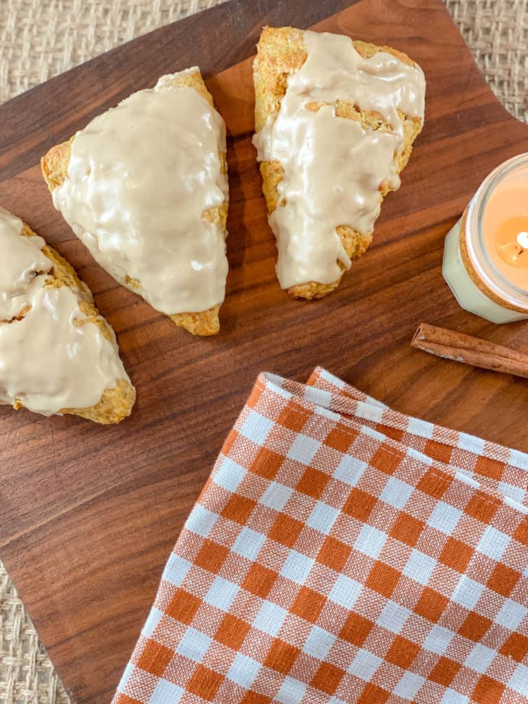 three pumpkin sourdough scones with maple glaze on bread board with candle, cinnamon stick, and orange plaid kitchen towel